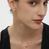 A woman with fair skin and brown hair pulled back wears a black top, a delicate gold necklace, and Georgie Crystal Knife Edge Huggie Hoop Earrings in Gold. She gazes softly to the side with a neutral expression.