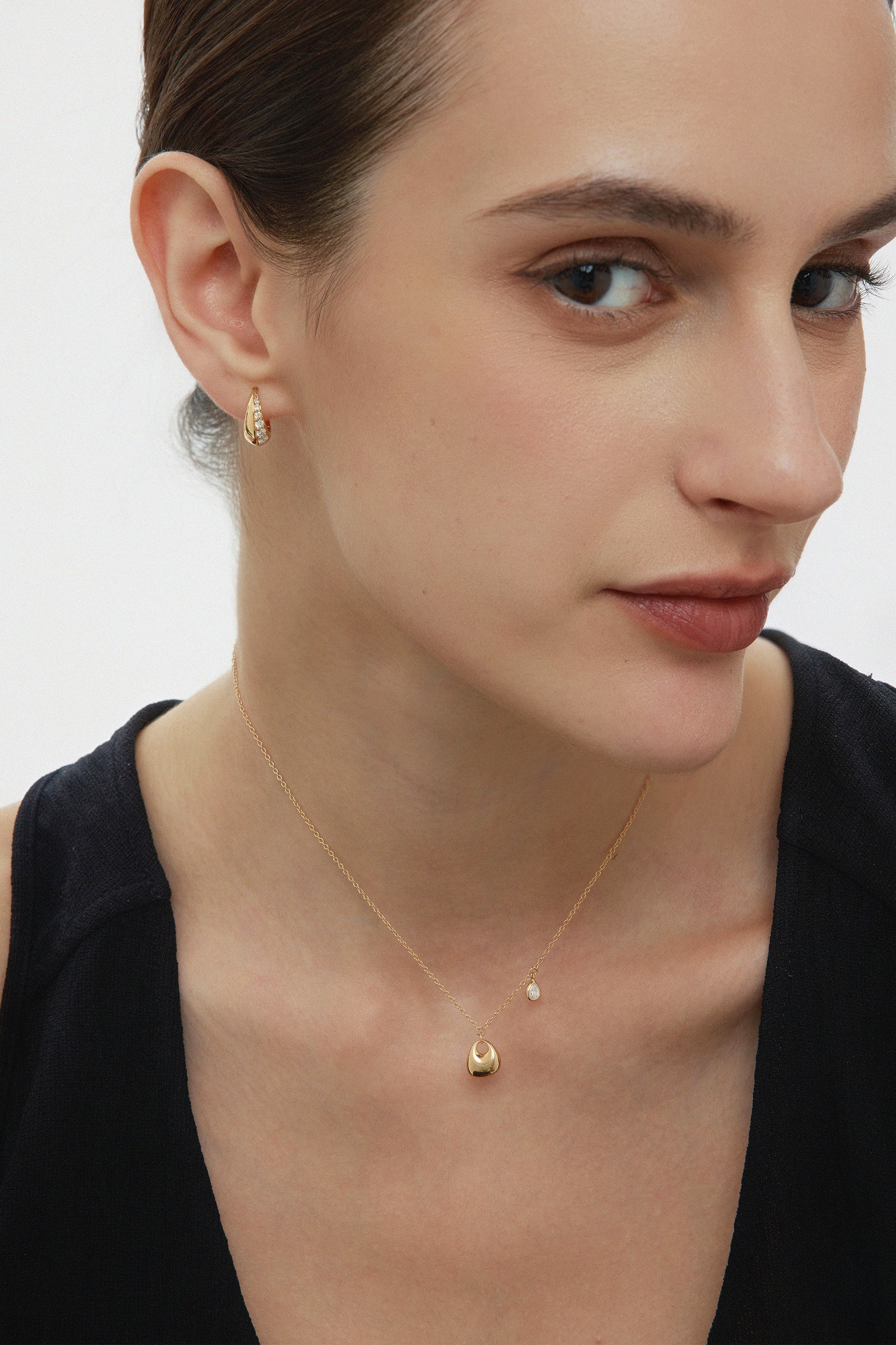 A woman with fair skin and brown hair pulled back wears a black top, a delicate gold necklace, and Georgie Crystal Knife Edge Huggie Hoop Earrings in Gold. She gazes softly to the side with a neutral expression.
