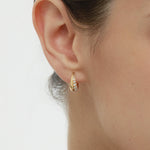 Close-up of a woman’s ear wearing the Georgie Crystal Knife Edge Huggie Hoop Earrings in Gold, featuring embedded cubic zircon stones. Her hair is tied back and a delicate gold chain necklace is partially visible.