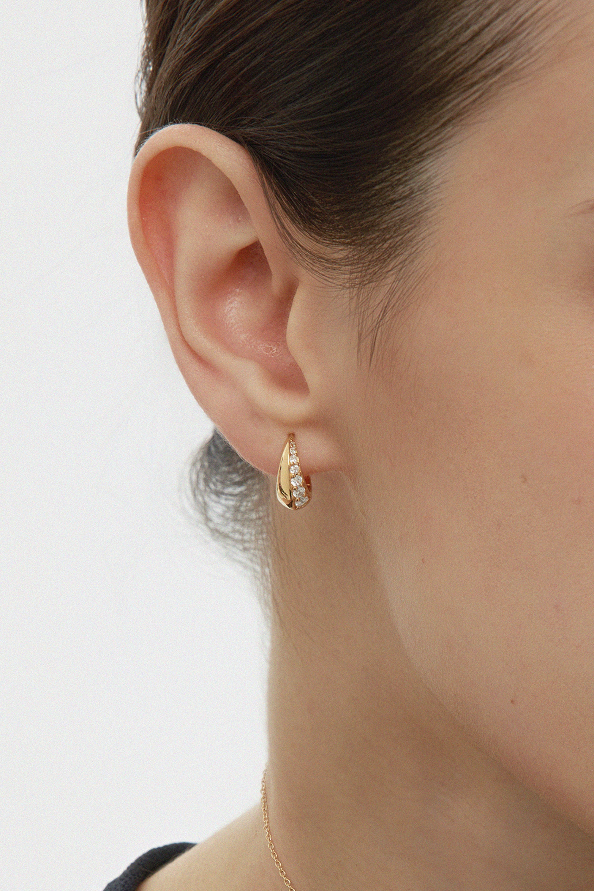 Close-up of a woman’s ear wearing the Georgie Crystal Knife Edge Huggie Hoop Earrings in Gold, featuring embedded cubic zircon stones. Her hair is tied back and a delicate gold chain necklace is partially visible.