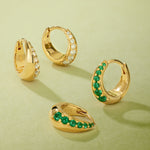 Four Georgie Crystal Knife Edge Huggie Hoop Earrings in Gold rest on a light green surface; two feature clear crystals, and two have green stones, each arranged at varying angles.