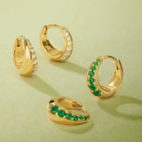 Four Georgie Crystal Knife Edge Huggie Hoop Earrings in Gold rest on a light green surface; two feature clear crystals, and two have green stones, each arranged at varying angles.