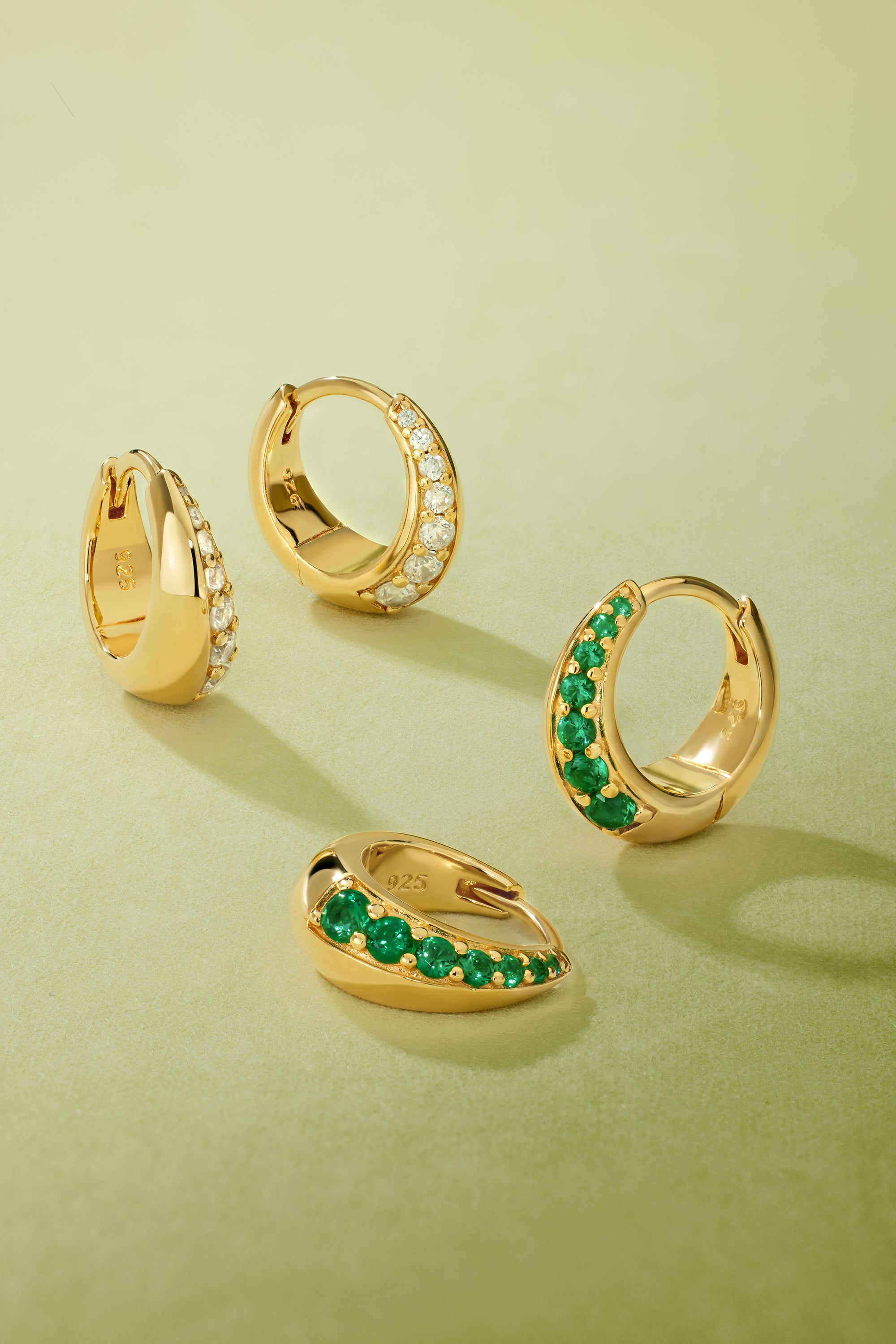 Four Georgie Crystal Knife Edge Huggie Hoop Earrings in Gold rest on a light green surface; two feature clear crystals, and two have green stones, each arranged at varying angles.