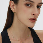 A woman with straight brown hair wears a black top, a gold necklace with green stones, and Georgie Crystal Knife Edge Huggie Hoop Earrings in Green, posing against a plain light background.