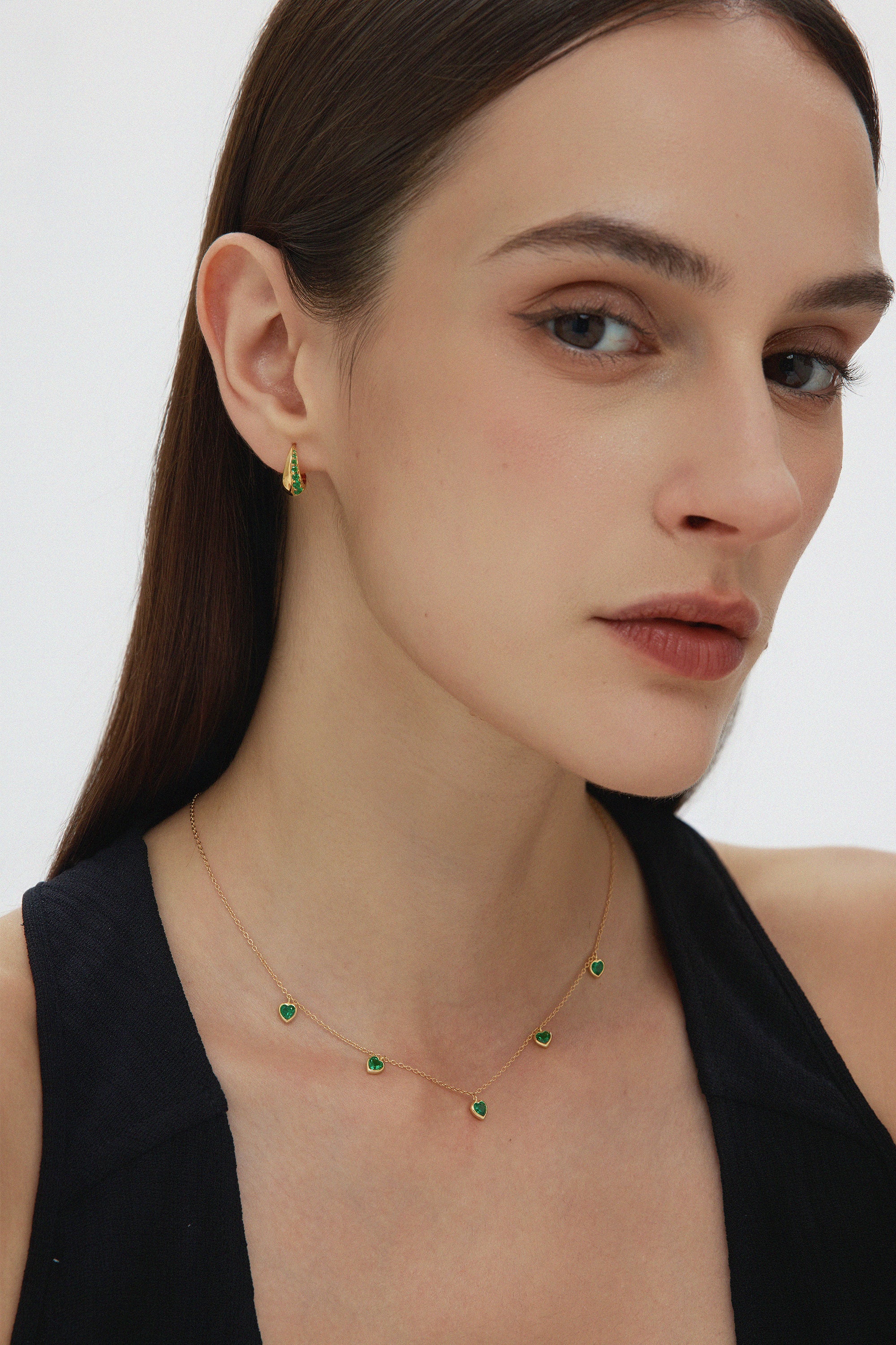 A woman with straight brown hair wears a black top, a gold necklace with green stones, and Georgie Crystal Knife Edge Huggie Hoop Earrings in Green, posing against a plain light background.