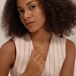 A woman with curly hair in a sleeveless, light-striped vest gazes softly at the camera. She wears gold jewelry, including a cubic zirconia ring and the Gisèle Cuban Link Bar Bracelet in Gold, and has a calm, neutral expression.