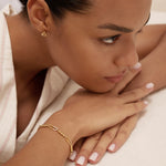 A woman with neatly styled hair and manicured nails gazes thoughtfully to the side, wearing the Carly Paperclip Chain Bead Bracelet in Gold with natural makeup and a heart-shaped earring.