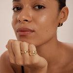 A woman with smooth skin and dark hair pulled back looks at the camera, wearing Colette Droop Link Huggie Earrings in Gold and gold rings, her chin resting on her hand.