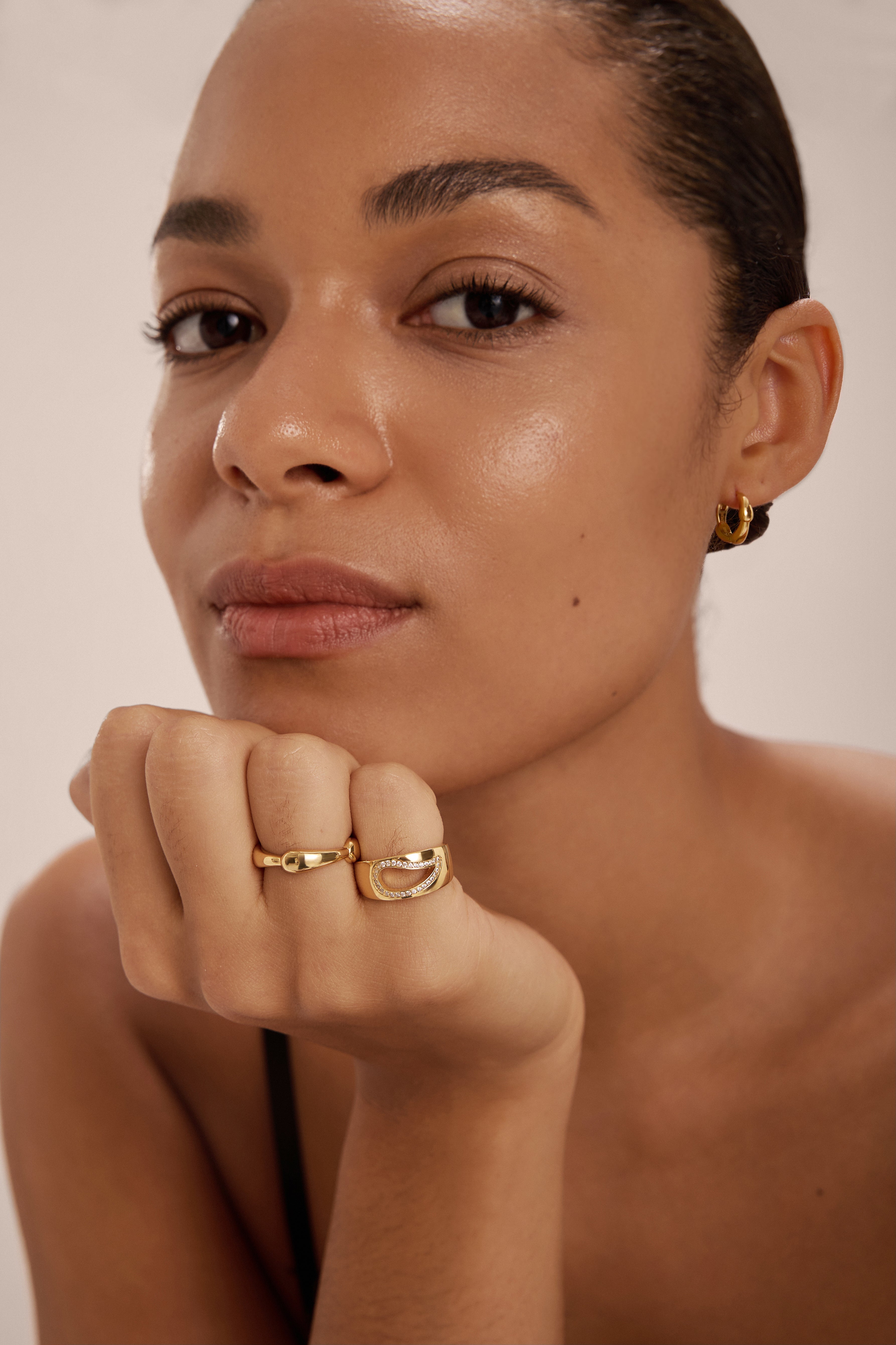 A woman with smooth skin and dark hair pulled back looks at the camera, wearing Colette Droop Link Huggie Earrings in Gold and gold rings, her chin resting on her hand.