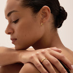 A woman with smooth, medium skin and dark hair in a low bun rests her hand on her shoulder, displaying the Colette Droop Link Huggie Earrings in Gold. She has neatly manicured nails, a peaceful expression, and an effortlessly elegant style.