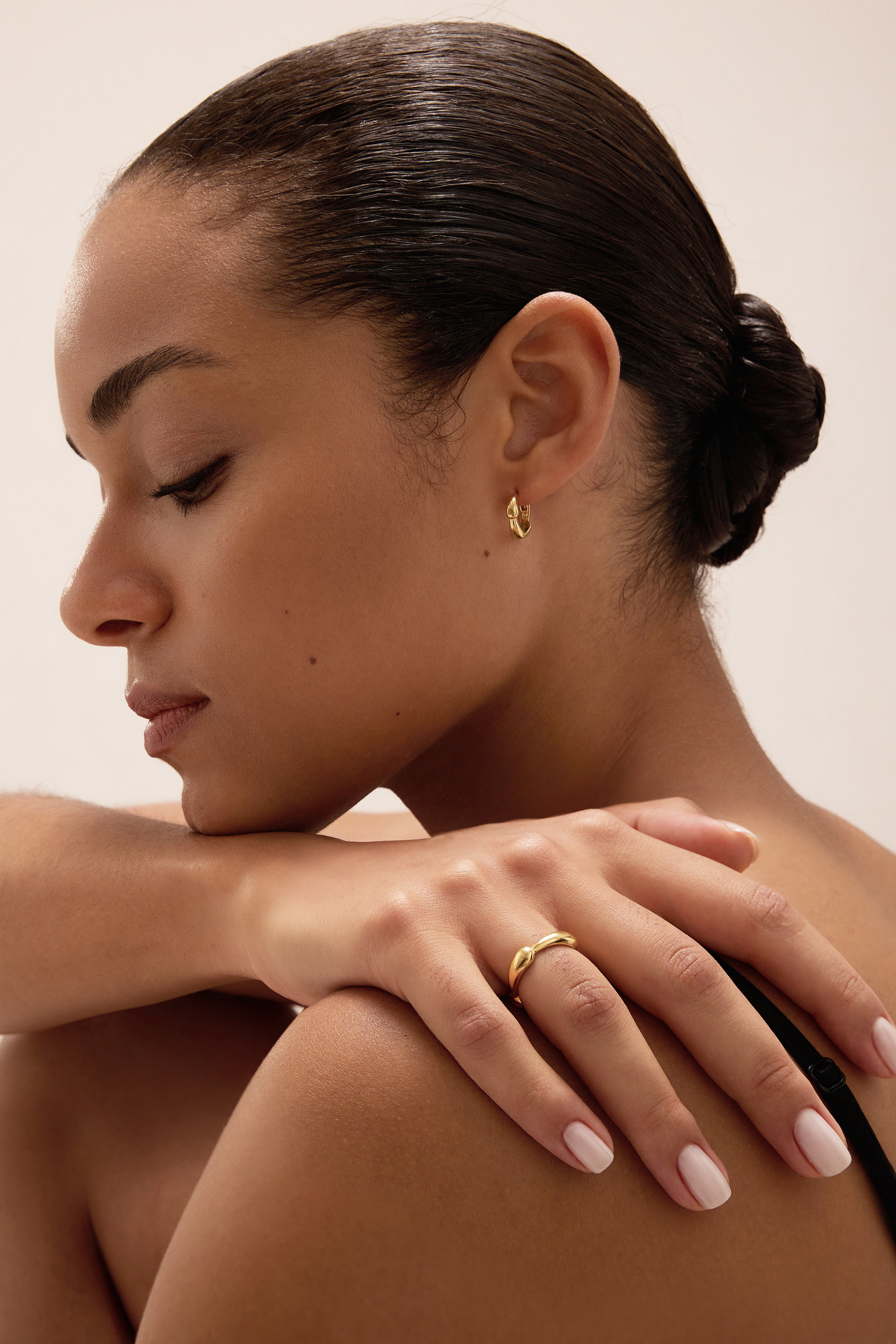 A woman with smooth, medium skin and dark hair in a low bun rests her hand on her shoulder, displaying the Colette Droop Link Huggie Earrings in Gold. She has neatly manicured nails, a peaceful expression, and an effortlessly elegant style.