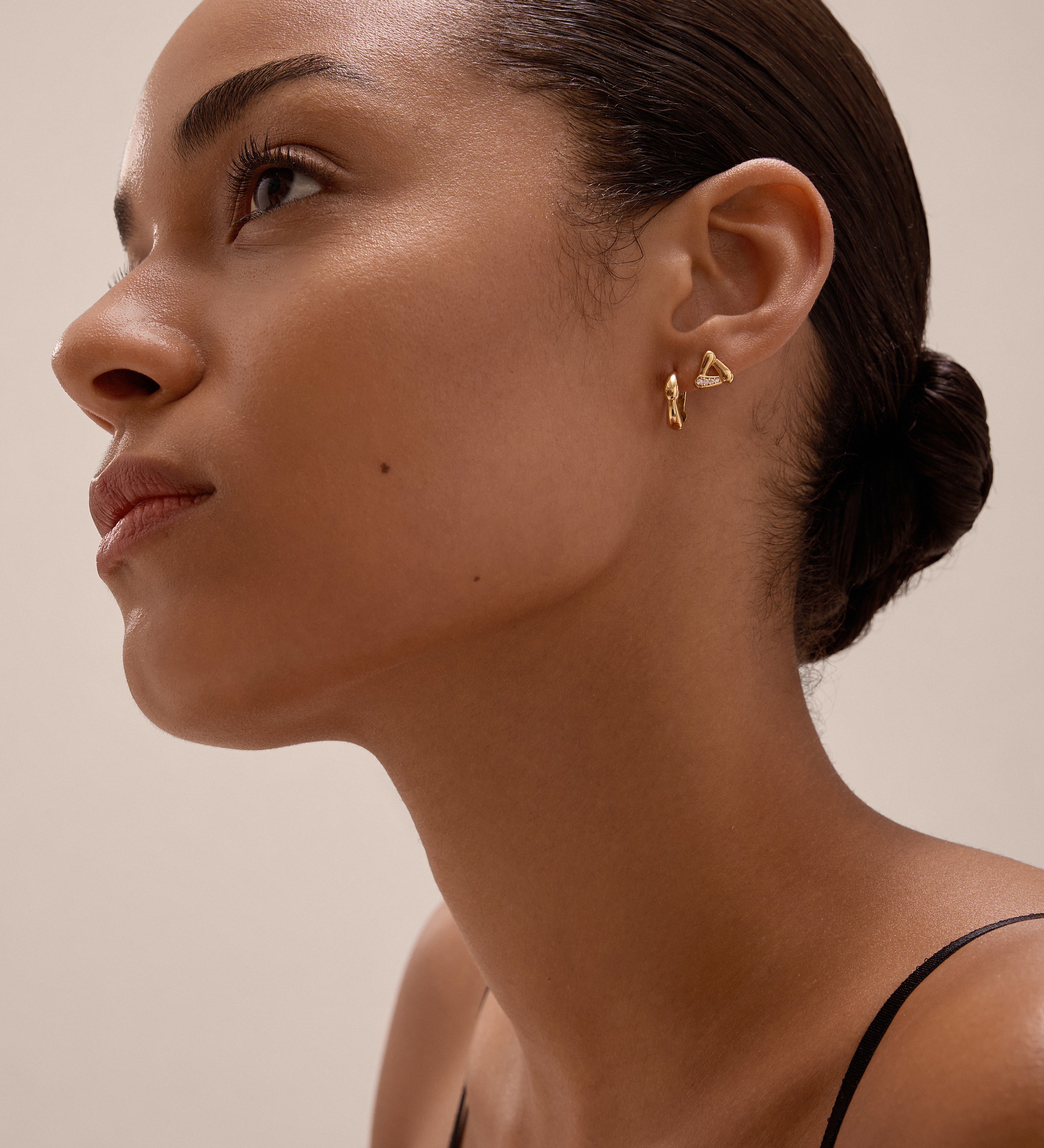 A woman with smooth, medium-brown skin and dark hair in a low bun gazes upward, wearing Colette Droop Link Studs in Gold and a black spaghetti strap top against a neutral background.