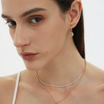 A woman with straight brown hair and natural makeup wears a white thin-strapped top and showcases the Crescent Moon Crystal Charm Huggies in Gold, layered with two other necklaces, against a plain background.