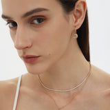 A woman with straight brown hair and natural makeup wears a white thin-strapped top and showcases the Crescent Moon Crystal Charm Huggies in Gold, layered with two other necklaces, against a plain background.