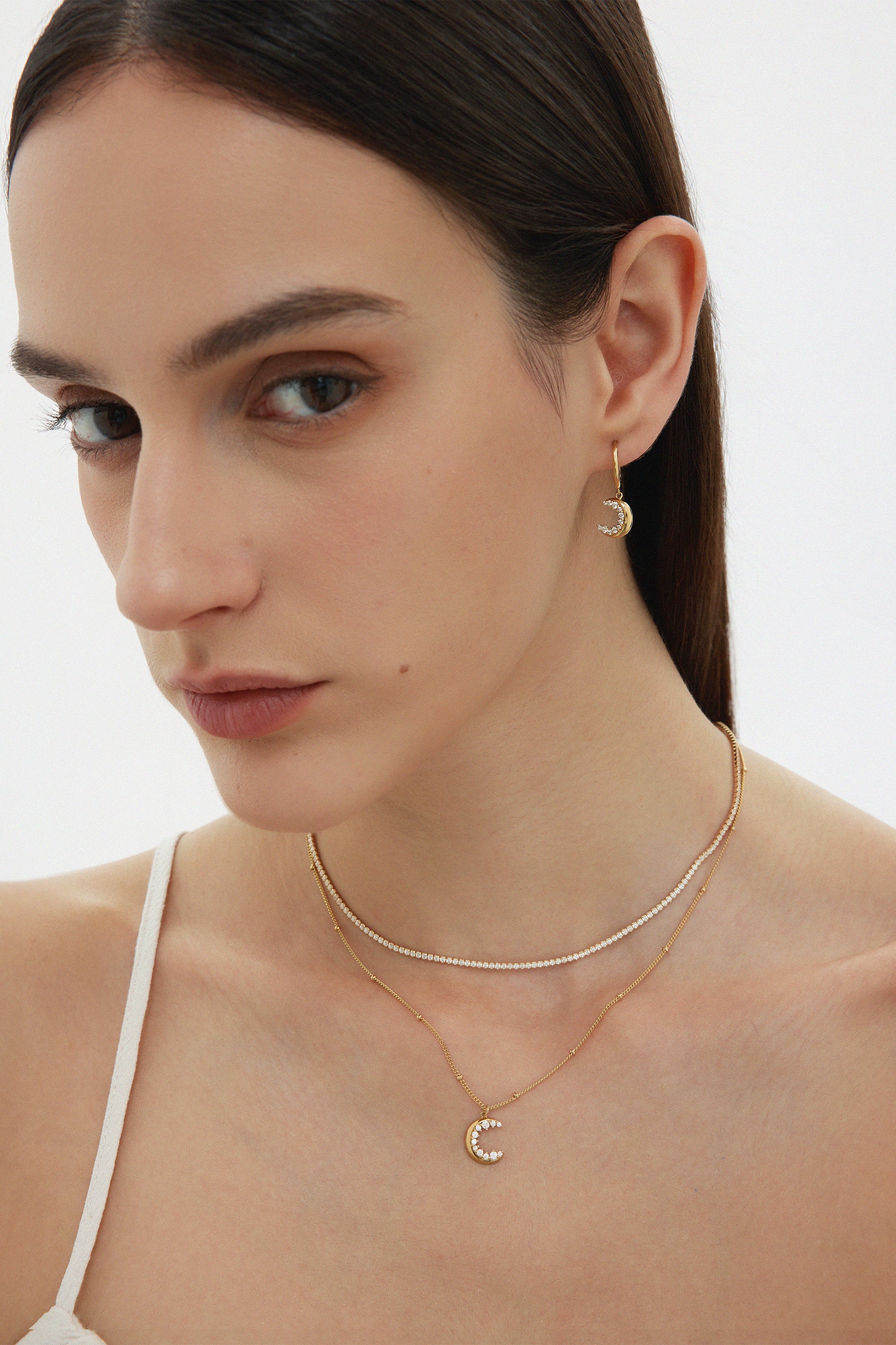 A woman with straight brown hair and natural makeup wears a white thin-strapped top and showcases the Crescent Moon Crystal Charm Huggies in Gold, layered with two other necklaces, against a plain background.