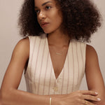 A woman with curly hair in a sleeveless, light top sits at a white table, looking to the side. She has a calm expression and wears delicate cubic zirconia jewelry and the Gisèle Cuban Link Bar Bracelet in Gold.