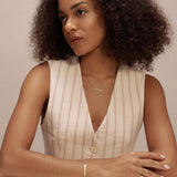 A woman with curly hair in a sleeveless, light top sits at a white table, looking to the side. She has a calm expression and wears delicate cubic zirconia jewelry and the Gisèle Cuban Link Bar Bracelet in Gold.