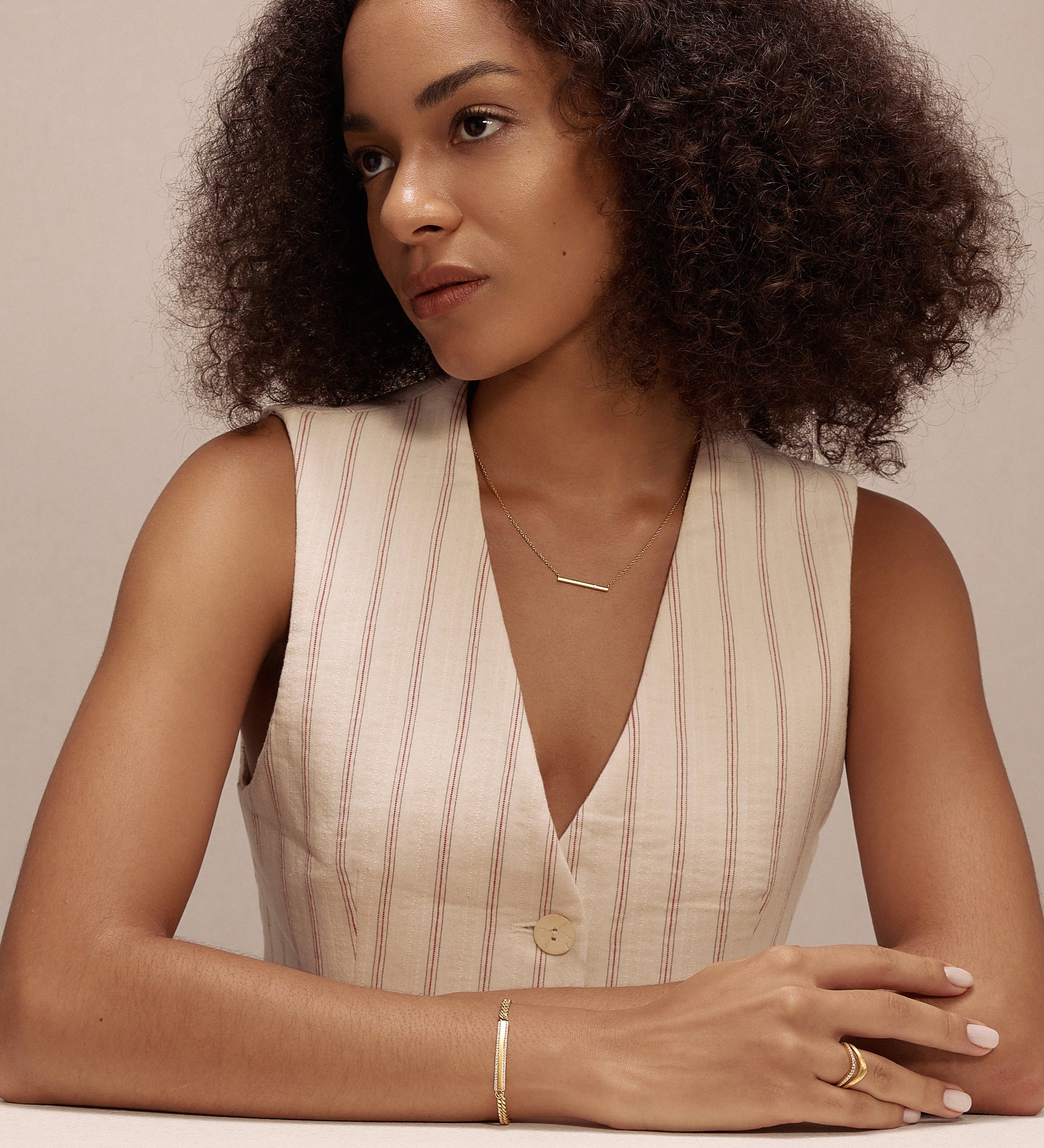 A woman with curly hair in a sleeveless, light top sits at a white table, looking to the side. She has a calm expression and wears delicate cubic zirconia jewelry and the Gisèle Cuban Link Bar Bracelet in Gold.