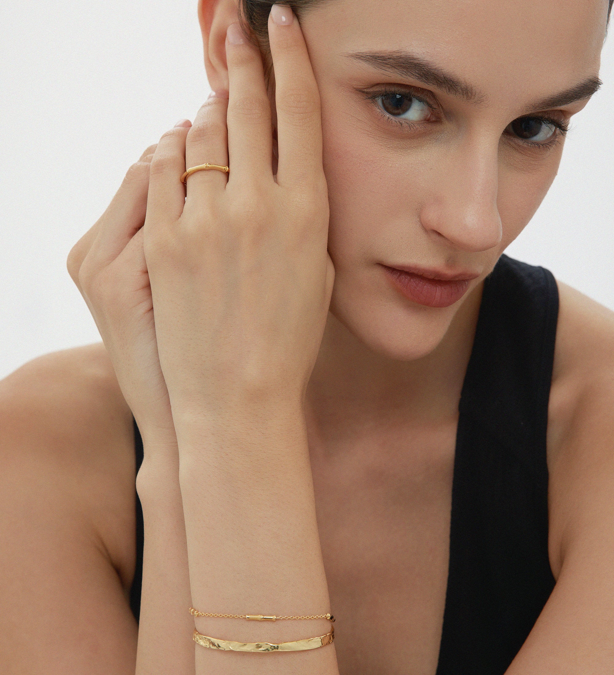 A woman with dark hair pulled back wears a sleeveless black top, a gold ring, and the Elodie Textured Torque Bangle in Gold, resting her hands on her face and looking at the camera against a plain background.