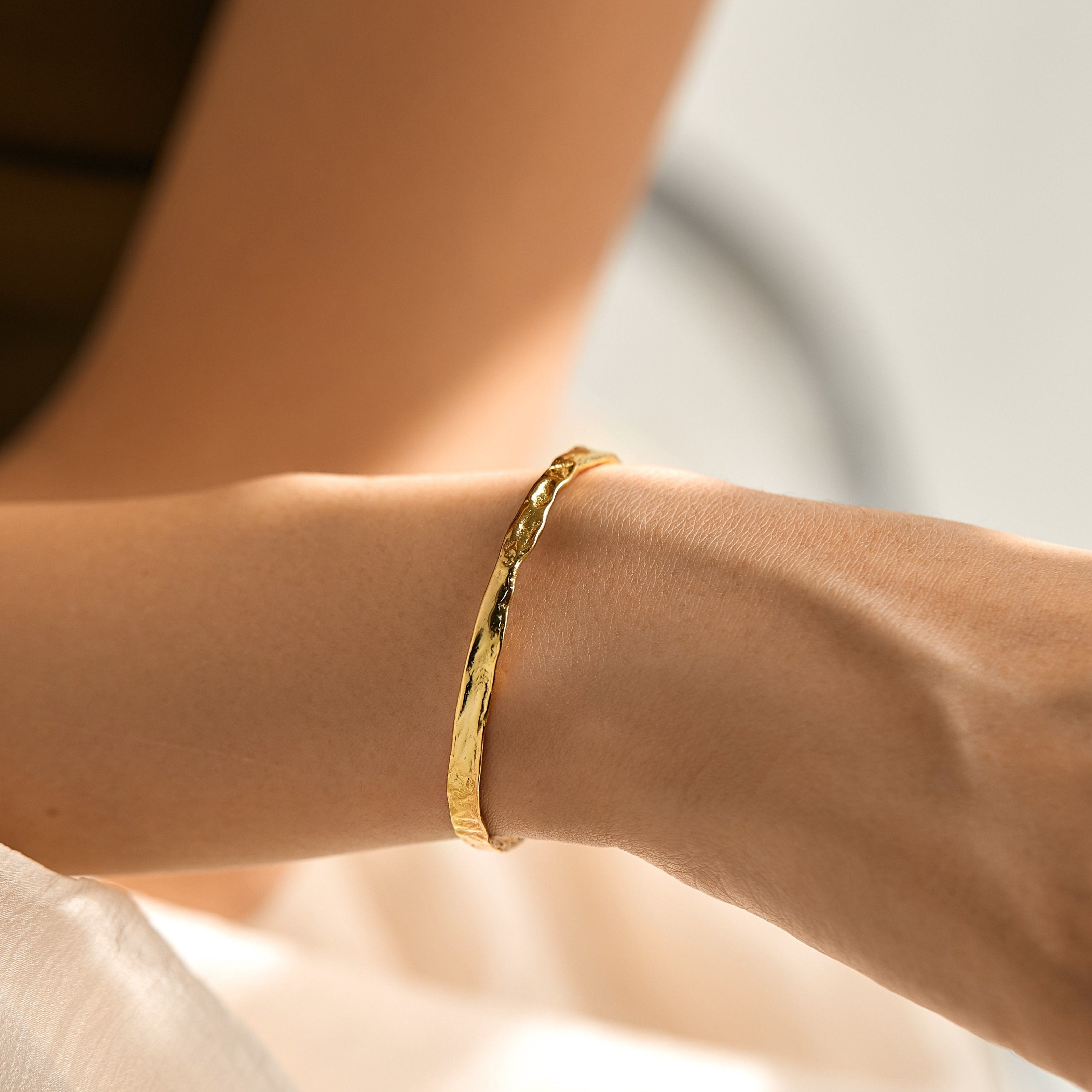 A close-up of a wrist wearing the Elodie Textured Torque Bangle in Gold, featuring a subtle lava-inspired texture. The softly blurred background enhances the bangle’s elegant shine and the smooth skin of the wearer.