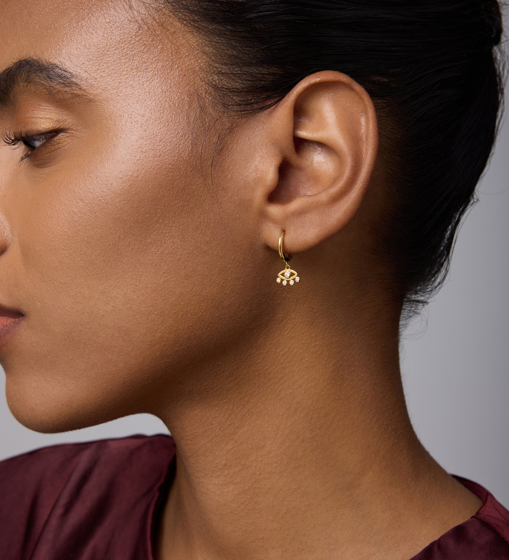 A woman with smooth skin and dark hair pulled back wears the Defender Evil Eye Hoops in Gold. She is dressed in a maroon top and shown in profile against a neutral background.
