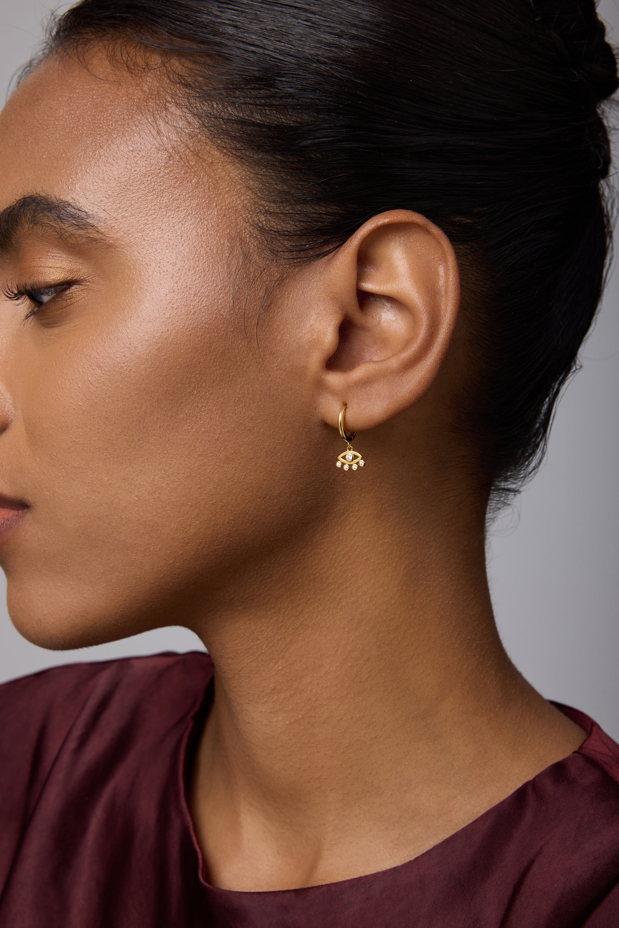 A woman with smooth skin and dark hair pulled back wears the Defender Evil Eye Hoops in Gold. She is dressed in a maroon top and shown in profile against a neutral background.
