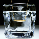 The Viva La Vida Bar Chain Necklace in Gold hangs over the rim of a clear glass filled with water, set against a white and black background, and features an adjustable figaro chain for the perfect fit.