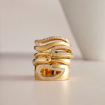 Three elegant gold rings, including the Colette Link Droop Ring in Gold with unique curves, are stacked together. The rings feature sparkling diamonds and one has a rectangular yellow gemstone. A blurred white object appears in the background.