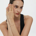 A woman with dark hair in a low bun wears a sleeveless black top and the Noor Pavé Triple Band Ring in Gold, resting her head on her hands and gazing thoughtfully to the side against a plain light background.
