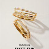 The Noor Pavé Triple Band Ring in Gold features overlapping bands and sparkling white gemstones, with one elegant pavé band. Shown on a light surface with shadows. Text below reads Featured in Vanity Fair.