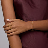 A person in a sleeveless burgundy satin top crosses their arms, displaying gold rings and the Pia Crystal Link Bracelet in Gold. Their neatly manicured nails are painted a light color.