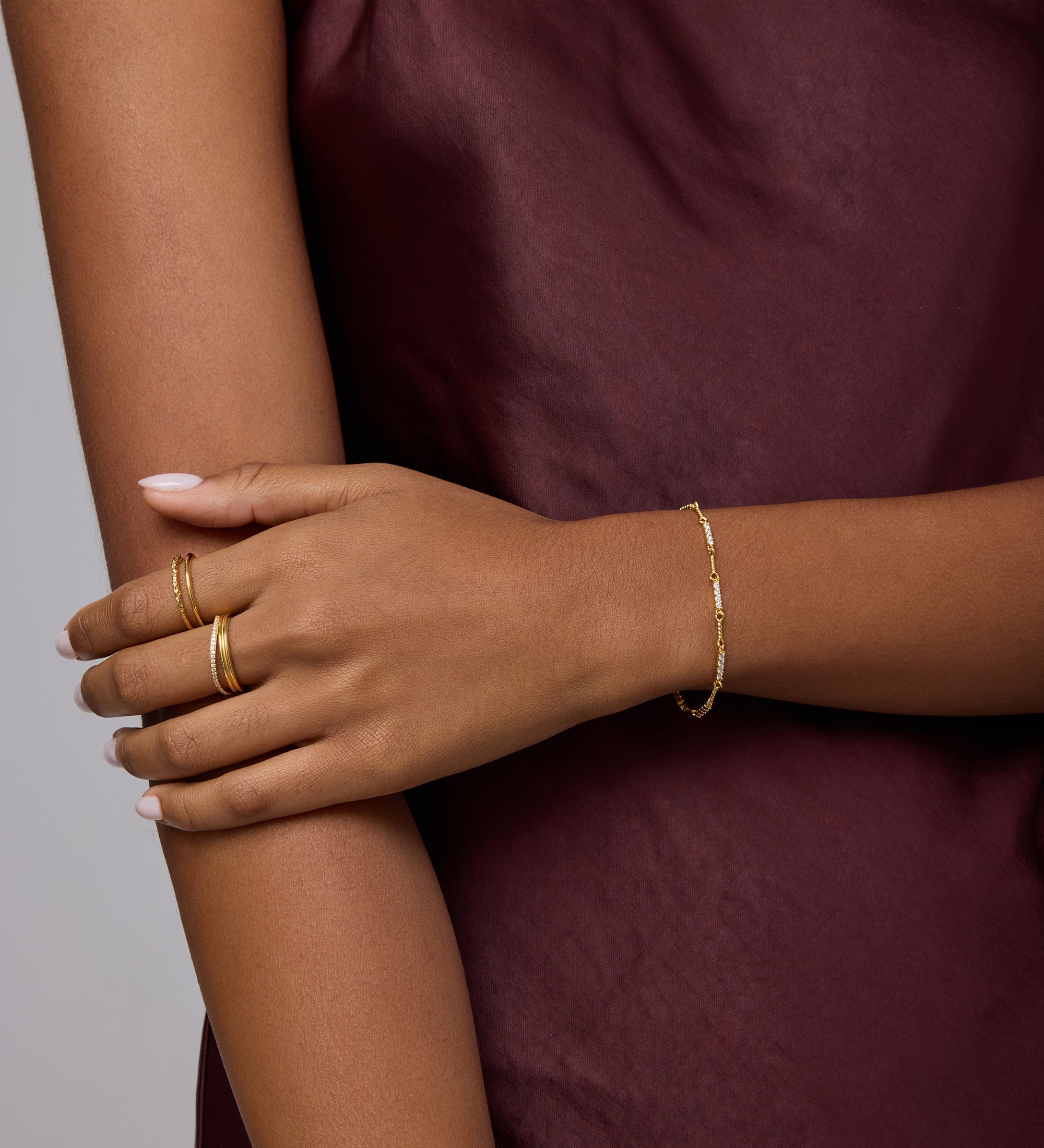 A person in a sleeveless burgundy satin top crosses their arms, displaying gold rings and the Pia Crystal Link Bracelet in Gold. Their neatly manicured nails are painted a light color.