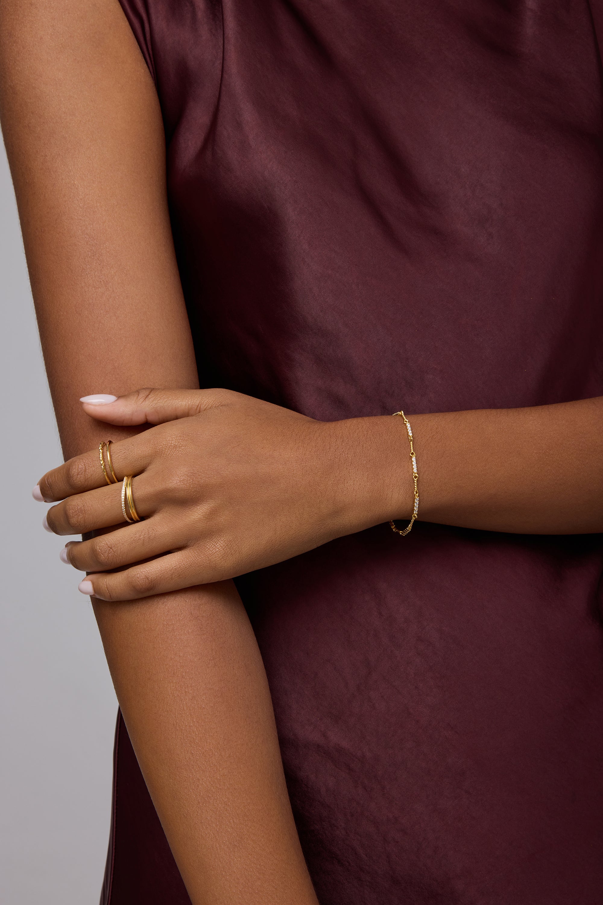 A person in a sleeveless burgundy satin top crosses their arms, displaying gold rings and the Pia Crystal Link Bracelet in Gold. Their neatly manicured nails are painted a light color.