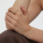 A person with light skin rests their hand on their arm, wearing the Noor Pavé Triple Band Ring in Gold and a delicate diamond bracelet. Theyre dressed in a dark brown garment against a plain, light-colored background.