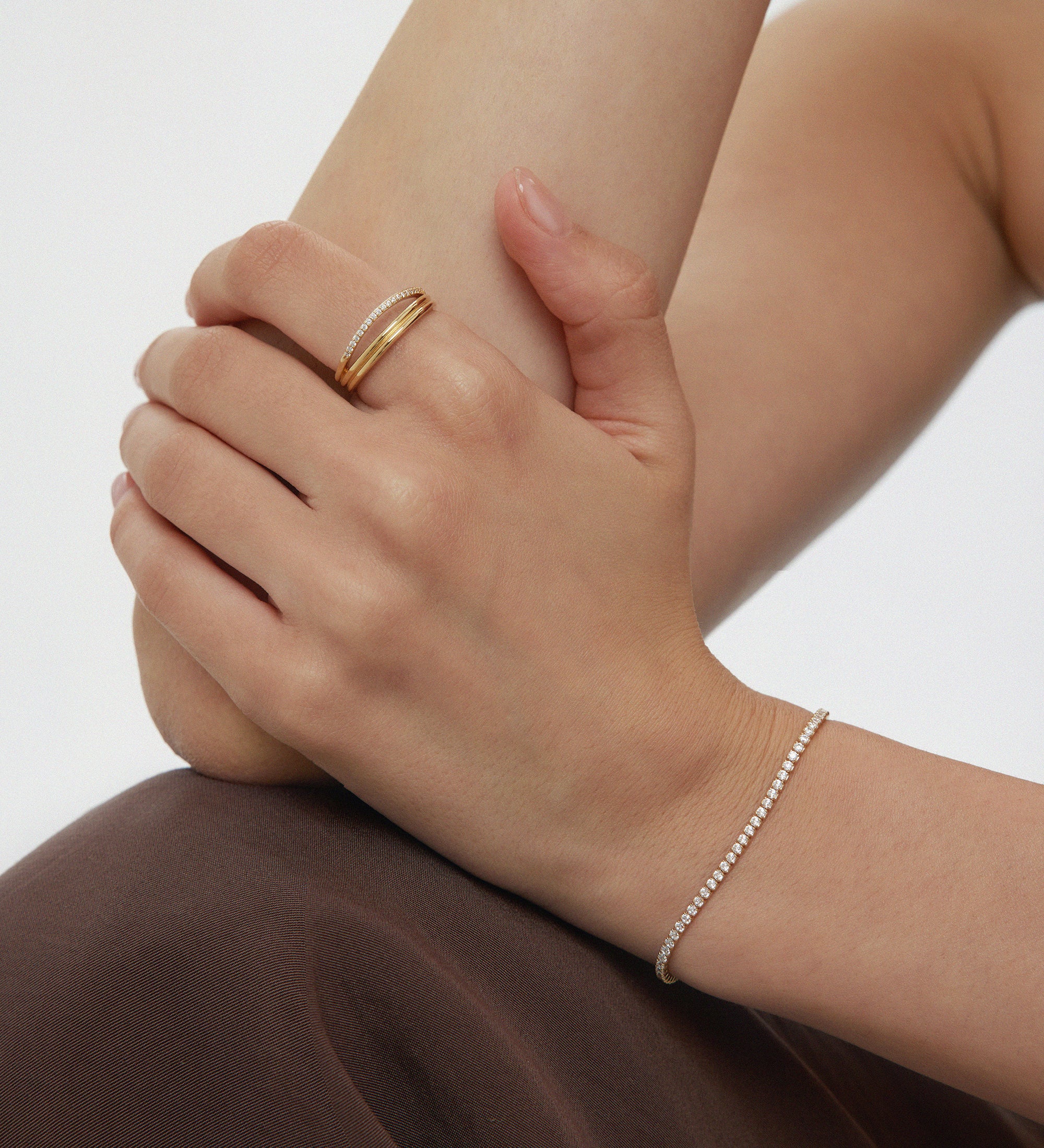 A person with light skin rests their hand on their arm, wearing the Noor Pavé Triple Band Ring in Gold and a delicate diamond bracelet. Theyre dressed in a dark brown garment against a plain, light-colored background.