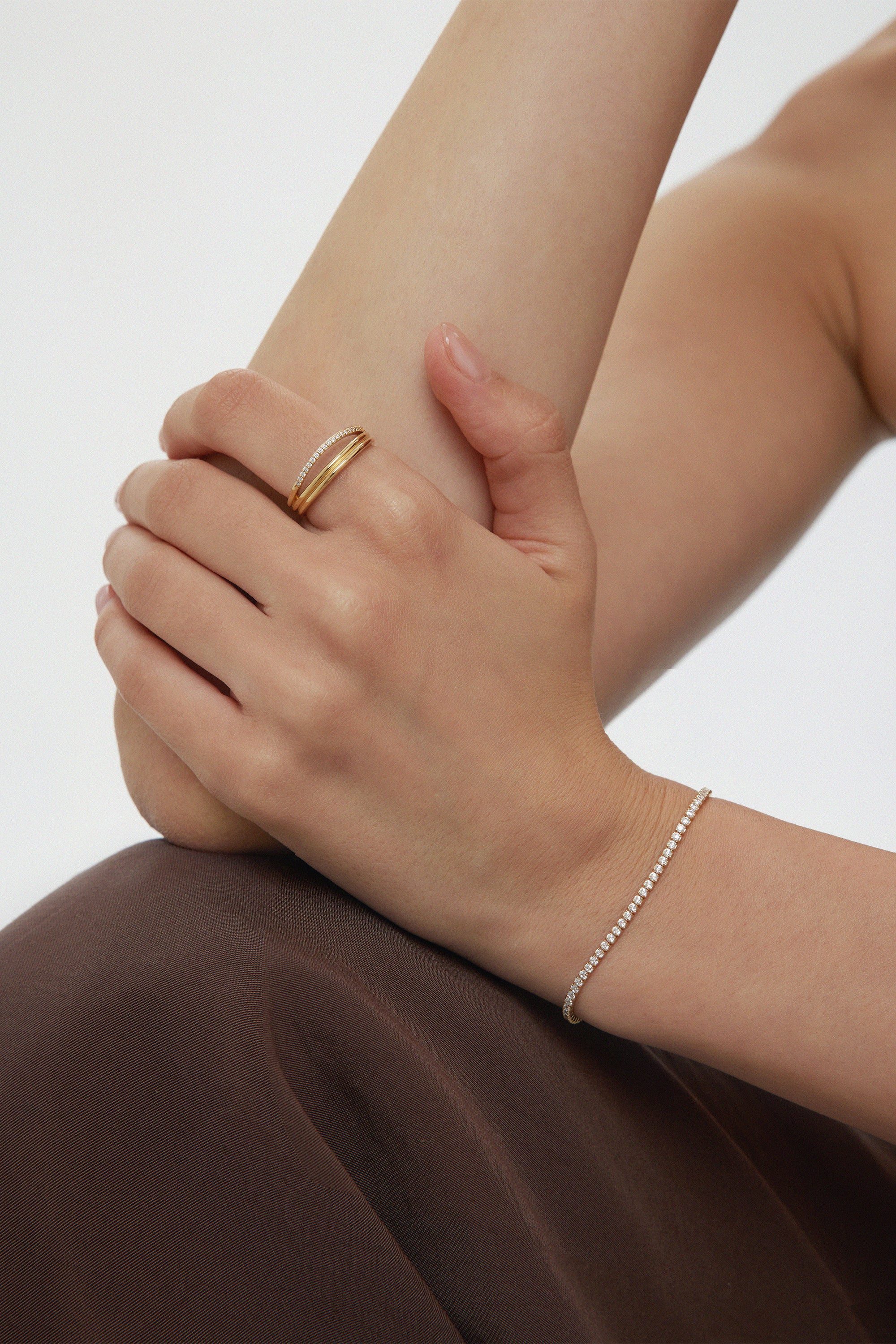 A person with light skin rests their hand on their arm, wearing the Noor Pavé Triple Band Ring in Gold and a delicate diamond bracelet. Theyre dressed in a dark brown garment against a plain, light-colored background.