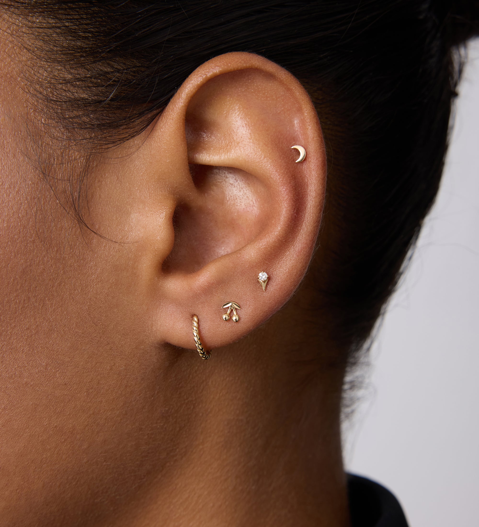 A close-up of an ear with four gold earrings: a crescent moon stud in the upper cartilage, 14K Recycled Solid Yellow Gold Rope Huggie Earrings in the lobe, and two small shaped studs above the huggie.