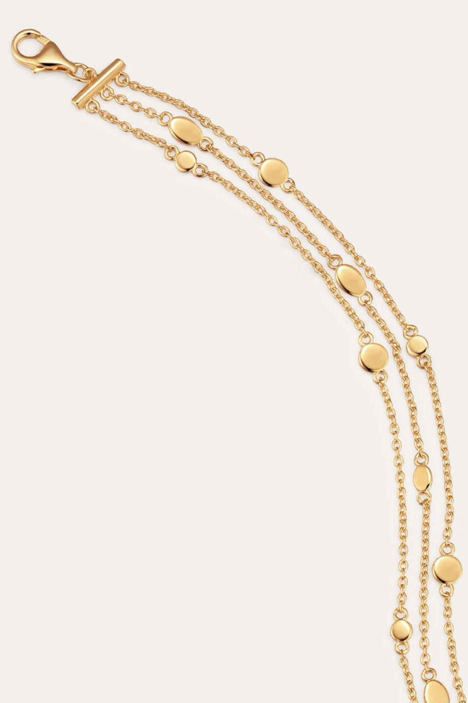 The Simone Triple Chain Layered Bracelet in Gold features three delicate 18k gold plated chains with small gold discs, secured by a toggle and lobster clasp, elegantly displayed on a plain light background.