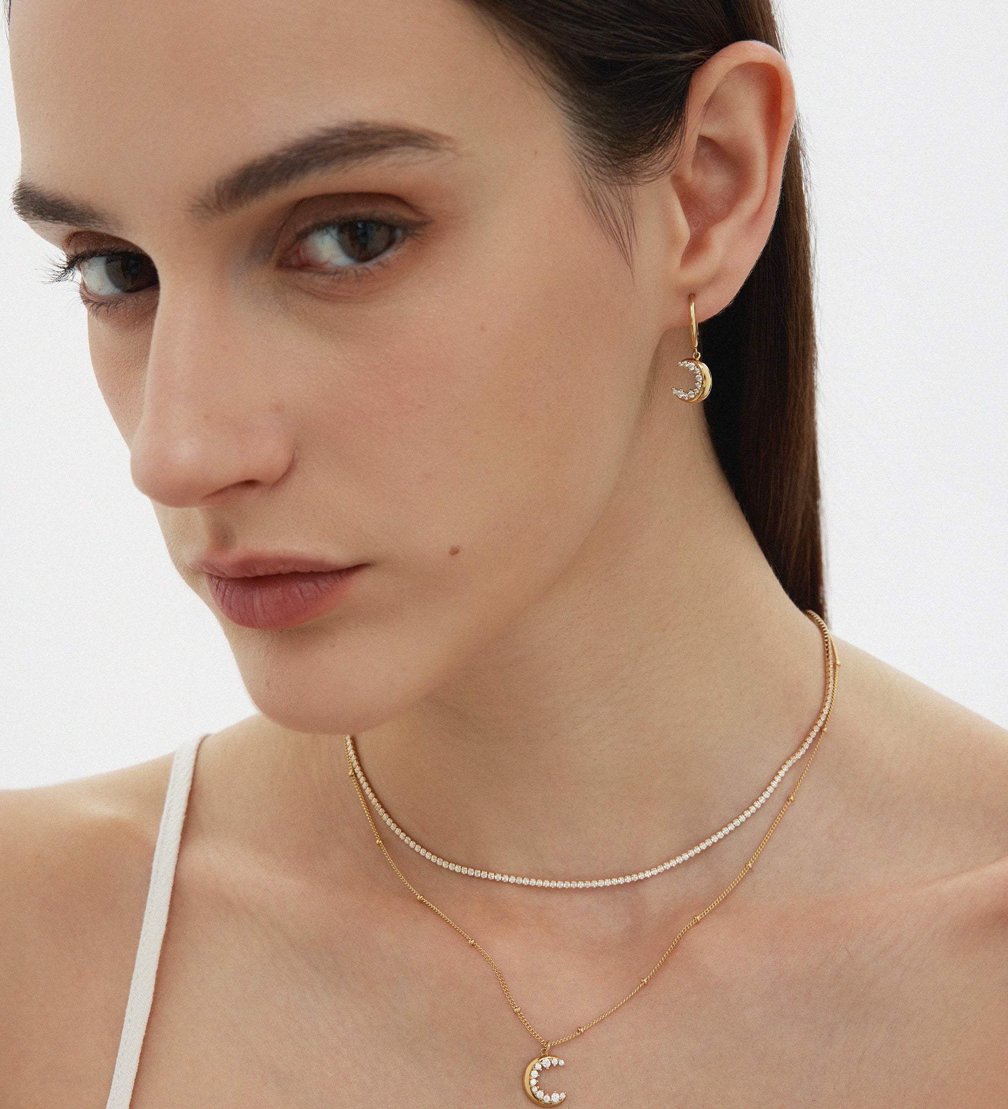 A woman with straight brown hair wears a white top, gold hoop earrings with a crescent moon charm, and a layered silver Alba Tennis Necklace—one strand features a matching crescent moon pendant. She has soft makeup and a neutral expression.