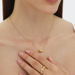 A woman with straight brown hair and natural makeup wears a gold necklace and the Vava Plain Chunky Curved Wave Ring in Gold. Her hand, adorned with the ring, rests gently on her chest as she gazes calmly ahead.