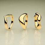 The Vava Plain Chunky Curved Wave Ring in Gold, with its modern sculptural form, stands upright on a reflective surface against a green-to-beige gradient background—a striking showcase of minimalist jewelry.