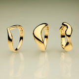 The Vava Plain Chunky Curved Wave Ring in Gold, with its modern sculptural form, stands upright on a reflective surface against a green-to-beige gradient background—a striking showcase of minimalist jewelry.