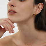 A woman with straight dark hair wears minimalist jewellery, featuring the Regina Horizontal Bar Pendant Necklace in Gold and small drop earrings, as she poses with her chin on her hand against a plain light background.