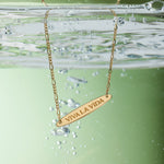 The Viva La Vida Bar Chain Necklace in Gold features VIVA LA VIDA engraved on a partially submerged pendant with bubbles and an adjustable figaro chain, set against a green background.