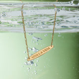 The Viva La Vida Bar Chain Necklace in Gold features VIVA LA VIDA engraved on a partially submerged pendant with bubbles and an adjustable figaro chain, set against a green background.