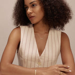 A woman with curly hair, wearing a sleeveless beige vest and the Regina Horizontal Bar Pendant Necklace in Gold, sits at a white table, arms resting on it, gazing thoughtfully to the side against a plain light background.
