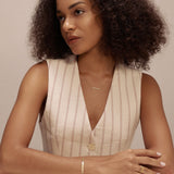 A woman with curly hair, wearing a sleeveless beige vest and the Regina Horizontal Bar Pendant Necklace in Gold, sits at a white table, arms resting on it, gazing thoughtfully to the side against a plain light background.