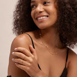A woman with curly hair wears a black sleeveless top and gold jewelry, including the Aisha Paperclip Stacking Ring in Gold. She smiles gently, looking to her right with her left hand resting on her right shoulder against a plain background.