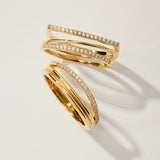 The Nola Pavé Triple Band Ring in Gold features three overlapping, pavé diamond-encrusted bands, elegantly displayed on a light beige surface with soft shadows.