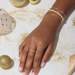 A hand adorned with the Celia Micro Freshwater Pearl Bracelet in Gold, along with gold and white rings, rests on a white surface scattered with gold glitter, ornaments, and a satin ribbon.